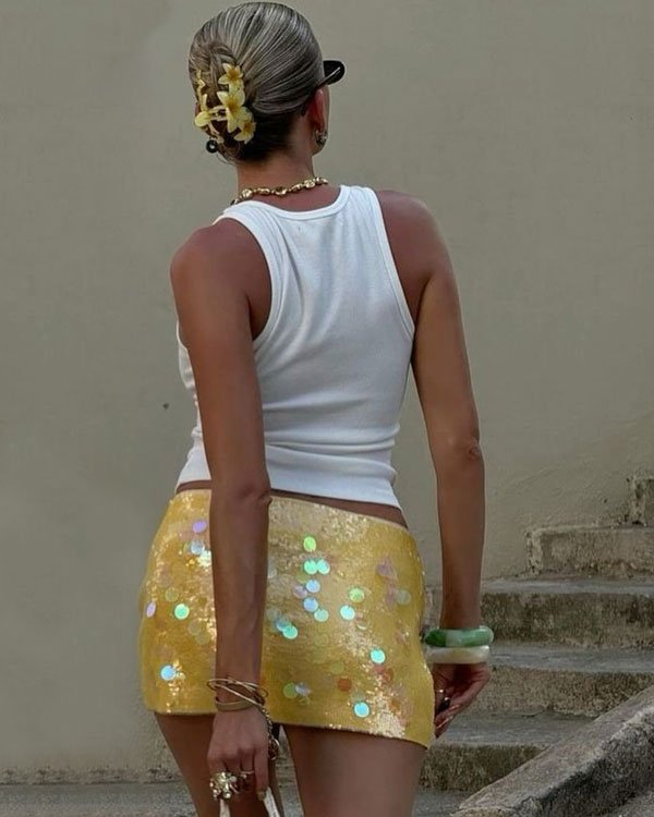 Mulher vestindo regata branca e saia curta dourada com lantejoulas iridescentes. Acessórios incluem pulseiras, anéis e um coque adornado com presilhas de flores. Look casual e moderno, ideal para clima quente e eventos informais.