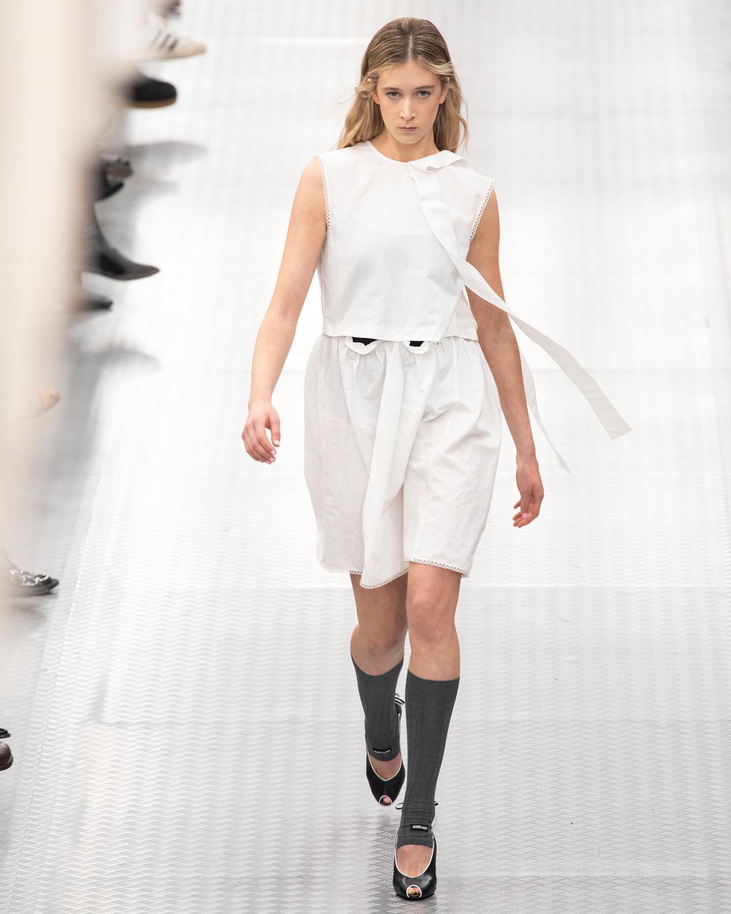 Modelo desfilando com vestido branco e meias cinza, refletindo tendências ousadas na moda.