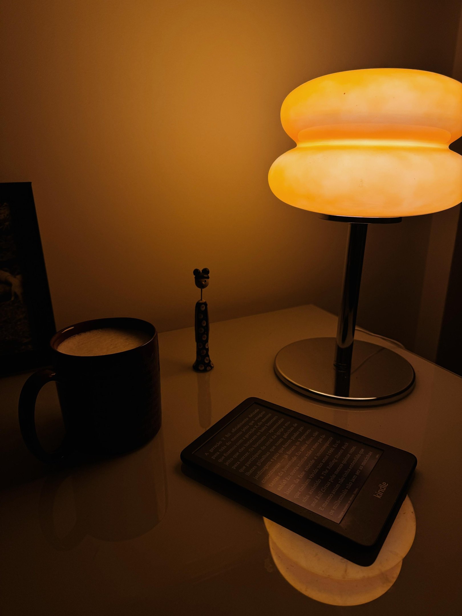 Imagem de um ambiente aconchegante, destacando uma luminária moderna com design curvilíneo e luz suave. Mesa de vidro com Kindle e xícara de café, evidenciando um estilo minimalista e tendências de simplicidade e conforto. Pessoa ansiosa.