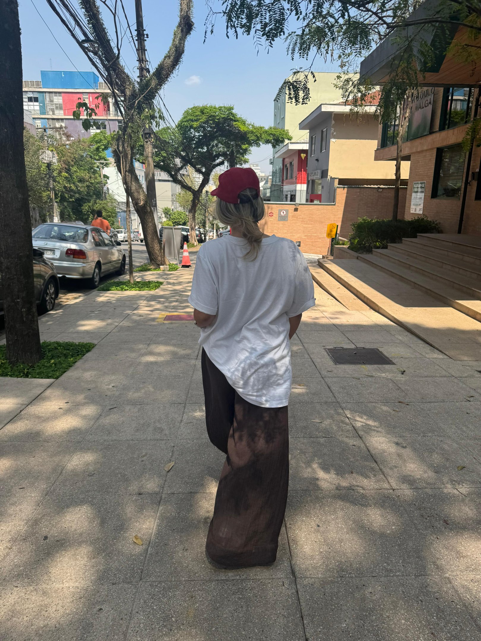 Pessoa caminhando pela cidade com estilo casual e descontraído, usando camiseta branca larga, calça marrom de tecido fluido e boné vermelho. Visual urbano, confortável e contemporâneo, tendência de moda atual. Pessoa ansiosa.