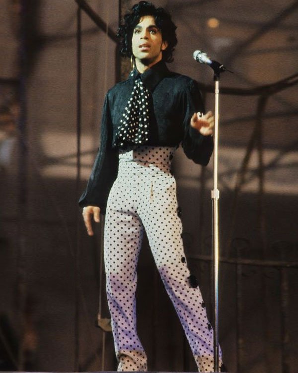 Artista em palco com blusa preta e calça de bolinhas, representando moda típica dos anos 80.