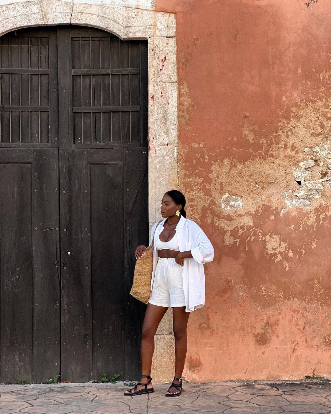 Mulher com look casual e elegante, usando conjunto branco de top, shorts e camisa leve, complementado por uma bolsa de palha e sandálias pretas. Estilo verão minimalista, com acessórios discretos e naturais. Rasteirinhas estilosas.