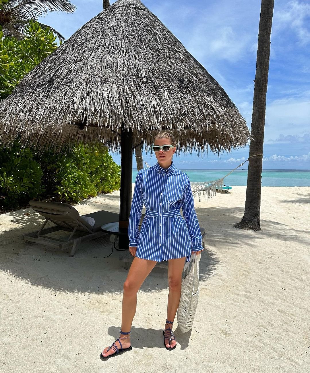 Pessoa em praia usando camisa de listras azuis e brancas como vestido, com cinto marcando a cintura. Sandálias pretas combinadas com óculos escuros brancos e bolsa de malha clara. Rasteirinhas estilosas.