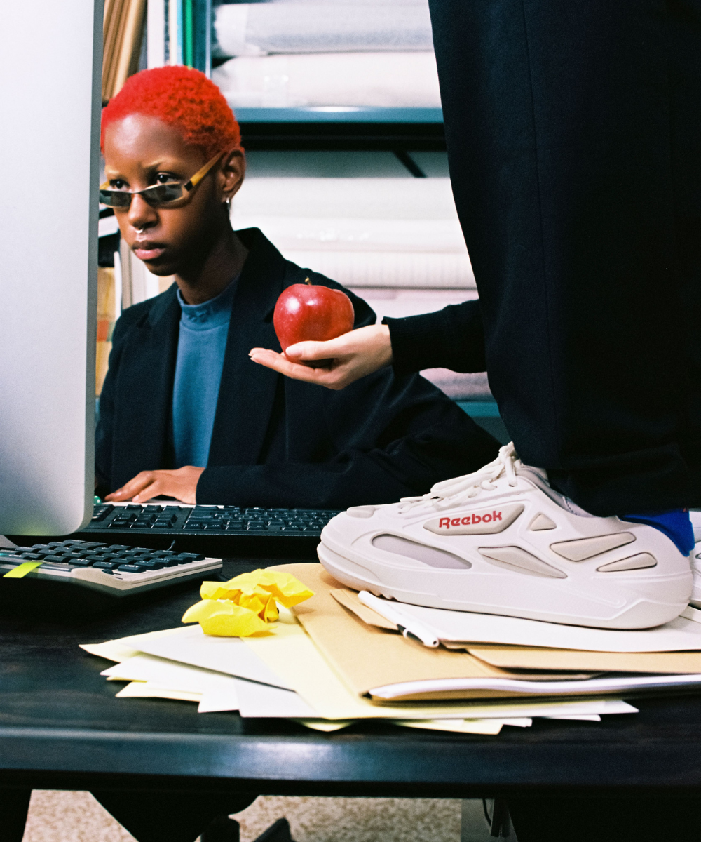 Pessoa com cabelo curto e tingido de vermelho, usando blazer largo preto sobre camiseta azul, óculos de sol pequenos e retangulares. Em segundo plano, uma mão segura uma maçã vermelha. Sobre a mesa, tênis branco Reebok volumoso em contraste com itens