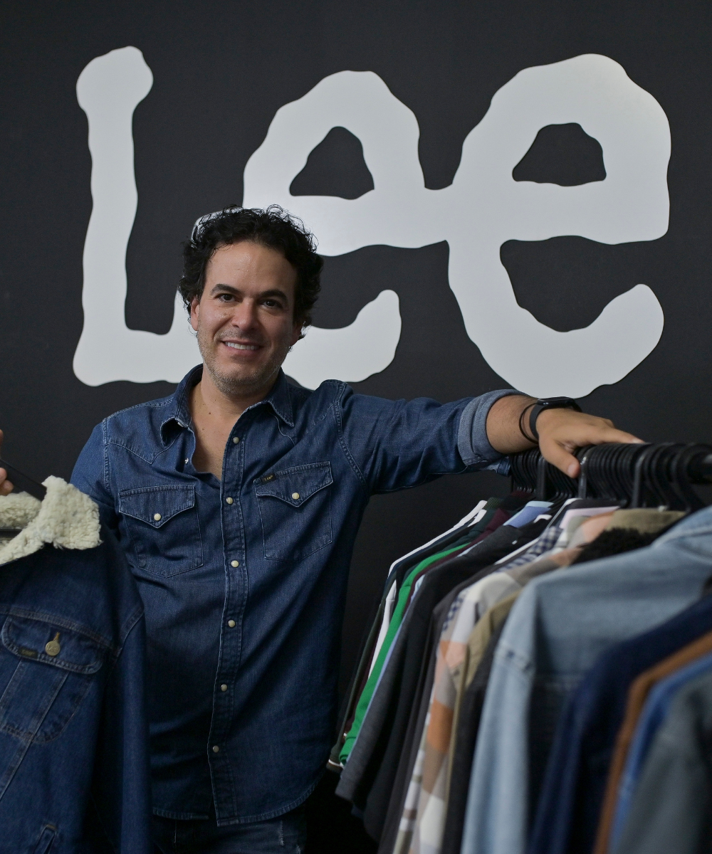 Homem vestindo camisa jeans posando ao lado de arara com roupas diversas. Estilo casual e atemporal, destacando tendências de moda denim. Fundo com logotipo da marca em destaque, criando um visual moderno e autêntico.