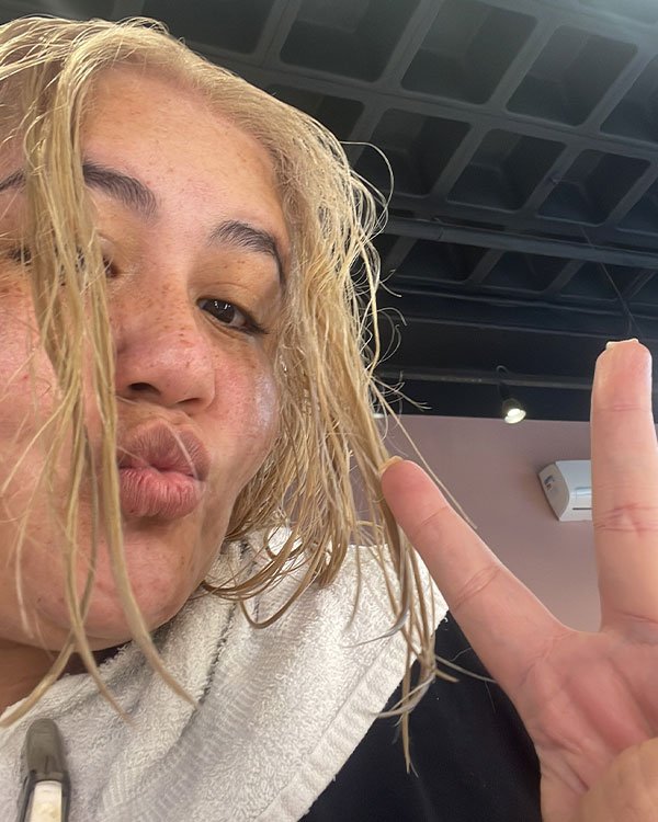 Pessoa com cabelo molhado loiro platinado, vestindo toalha branca no ombro. Estilo casual despojado, tendência de selfies descontraídas em ambientes internos. Sinal de paz com os dedos, popular em redes sociais.