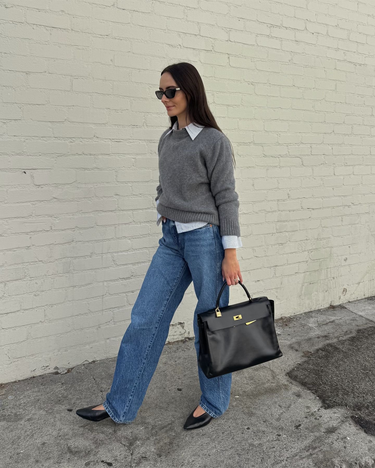 Mulher com suéter cinza e jeans, usando sapato clássico e elegante preto, caminha carregando bolsa preta.