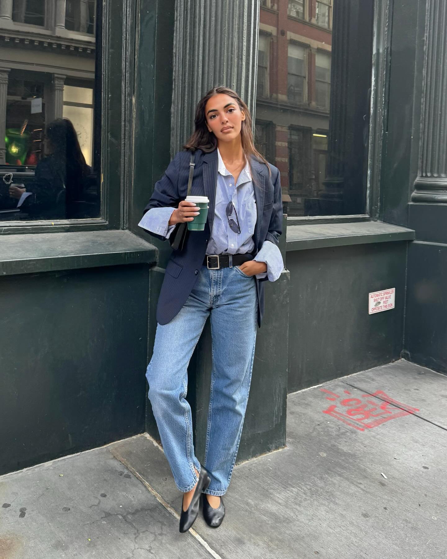 Mulher com camisa azul, blazer, calça jeans e sapato clássico elegante, segurando café.