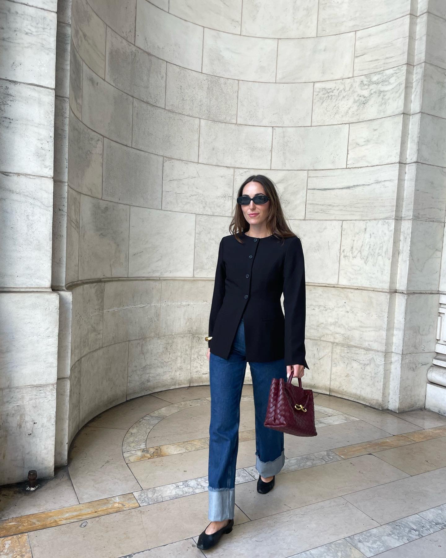 Mulher com blazer preto, jeans e sapato clássico e elegante, segura bolsa vinho e usa óculos de sol.