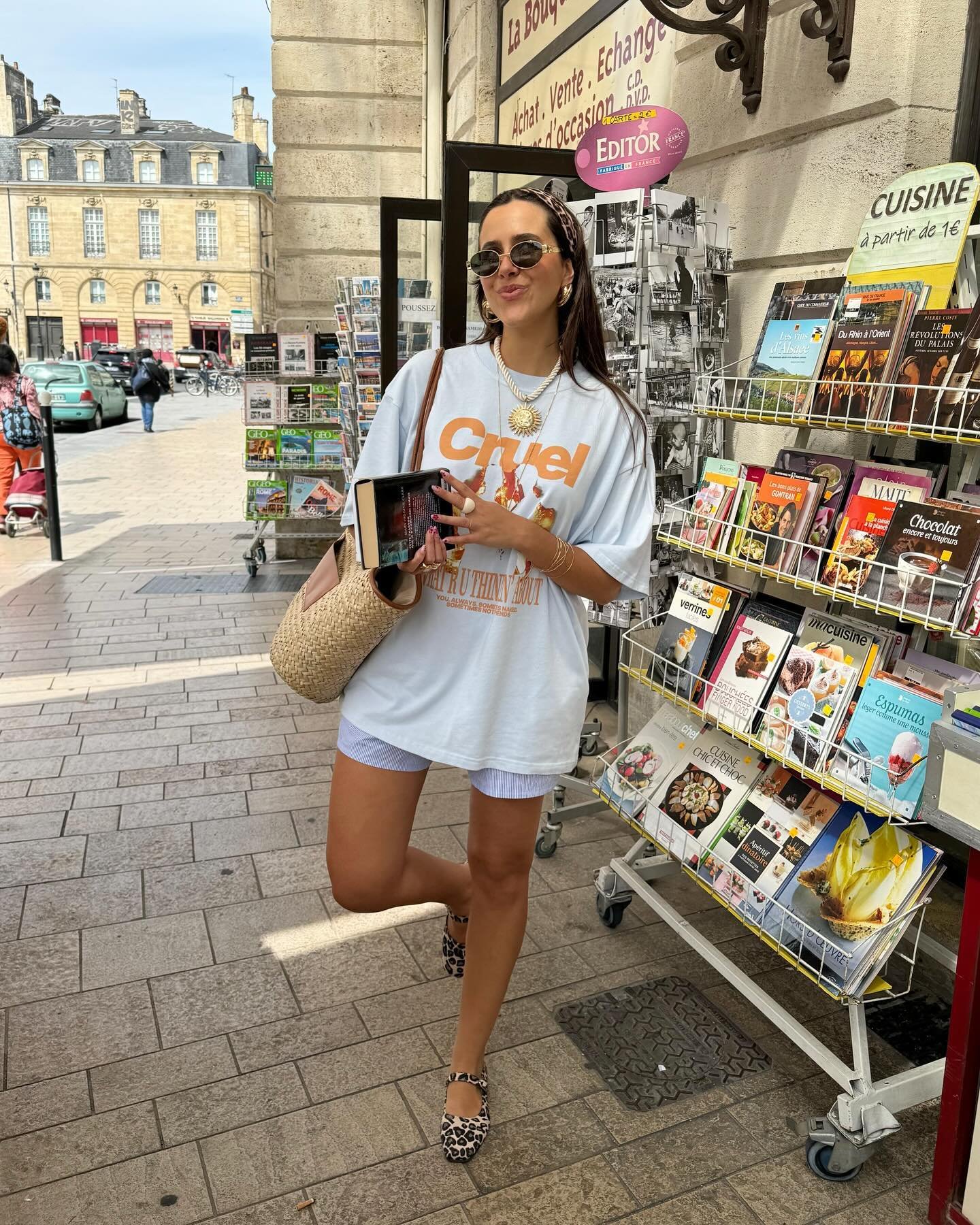 Pessoa com camiseta branca e sapatos chiques estampados, em frente a banca de revistas em rua movimentada.