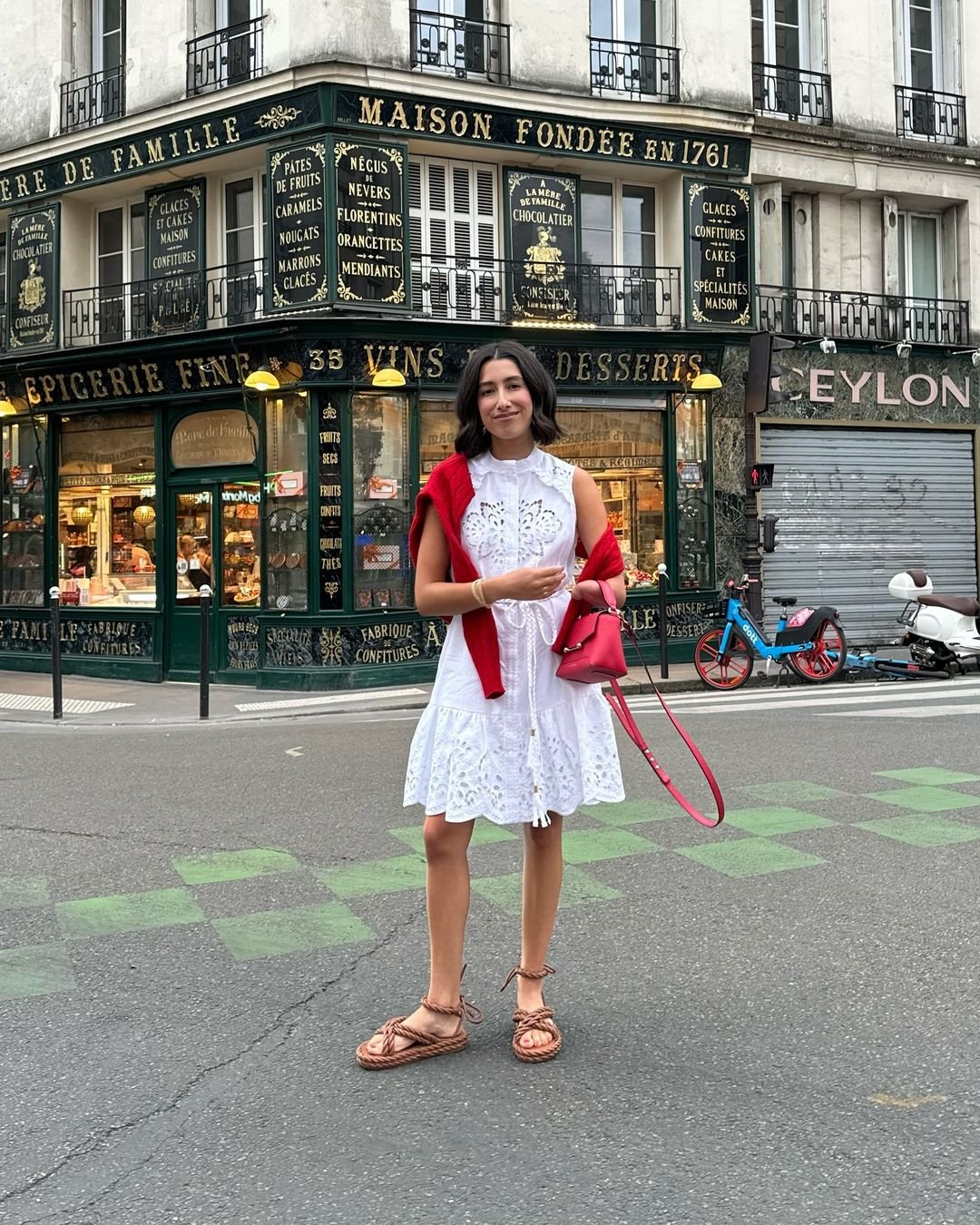 Mulher vestindo um vestido branco rendado com um suéter vermelho sobre os ombros e uma bolsa vermelha. Ela usa sapatos tendência. Estilo casual elegante, ideal para passeios urbanos. Fundo com arquitetura francesa tradicional.