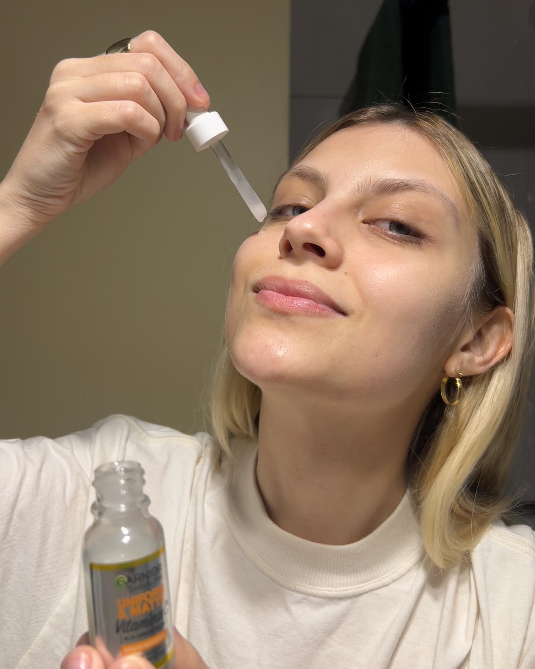 Mulher aplicando sérum com vitamina c facil. Ela usa blusa branca de gola alta e brincos de argola dourados, ilustrando tendências minimalistas e de autocuidado. A moda se destaca pelo foco em beleza natural e simplicidade.