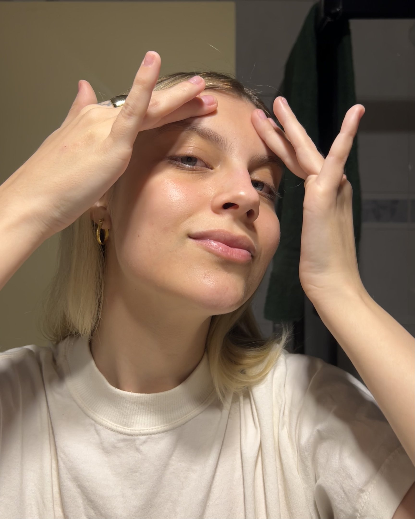 Moça em camiseta branca casual realiza cuidados faciais. O look clean e minimalista, marcado por acessórios discretos como brincos pequenos e anel robusto, reflete tendências contemporâneas de simplicidade e autocuidado.