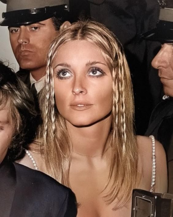 Sharon Tate com cabelos loiros compridos, destacando pequenas tranças finas. Vestido de alças finas e maquiagem suave, realçando os olhos. Estilo boêmio e elegante, refletindo a moda hippie-chic da época.