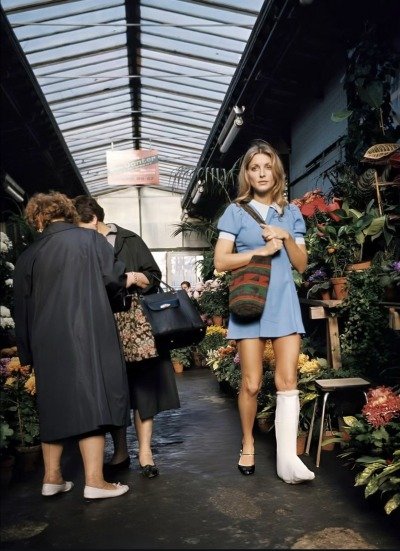 Uma mulher vestindo um vestido azul curto e moderno na moda dos anos 60, com uma bota ortopédica branca. Ela carrega uma bolsa de ombro listrada em um mercado de flores. Ao fundo, pessoas em casacos sóbrios observam as plantas.