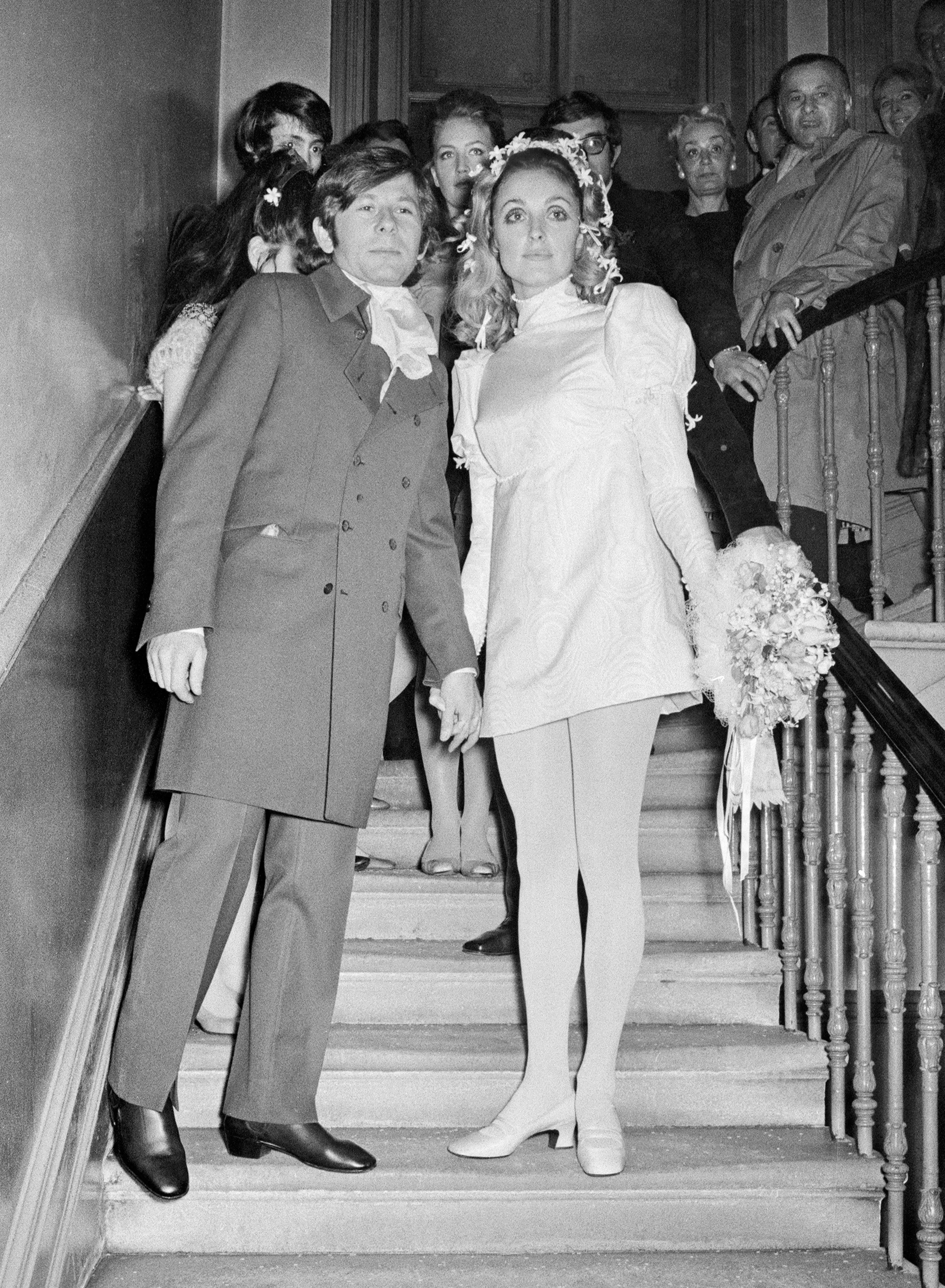 Casal em um casamento dos anos 60. Sharon Tate usa terno com casaco longo e calça ajustada, complementado por camisa volumosa. Ela veste minivestido branco com mangas longas, meia-calça e sapato de salto, com flores no cabelo. Estilo retrô e romântico.