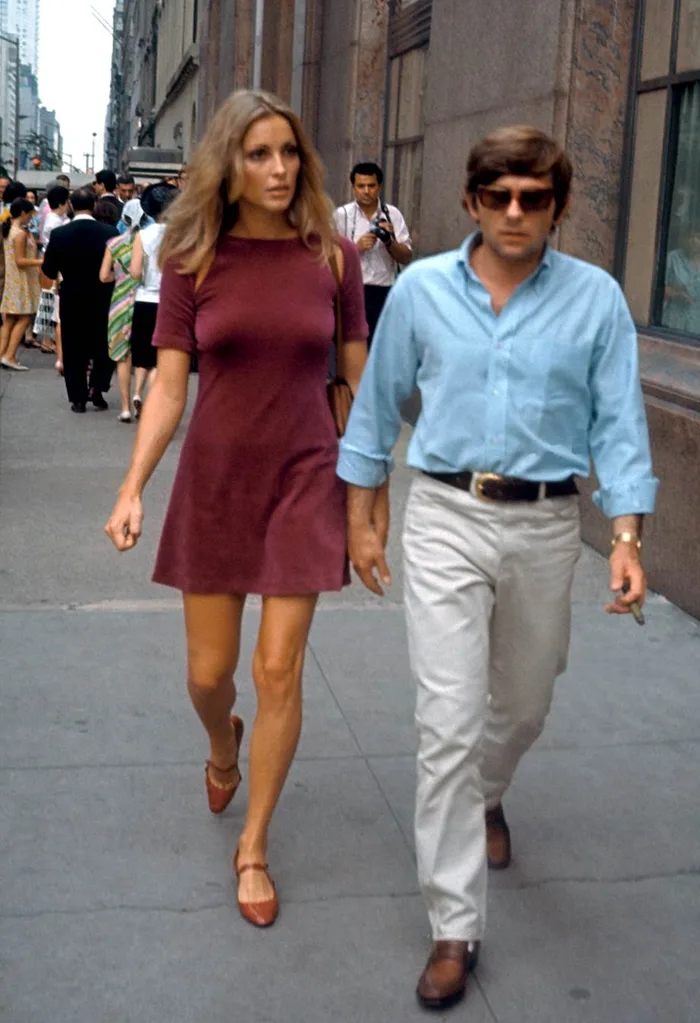 Casal caminhando em rua urbana dos anos 60. Ela usa vestido curto vinho e sapatos marrons, estilo mod. Ele veste camisa azul clara e calças claras, com cinto marrom. Ambos apresentam moda clássica e casual da década.