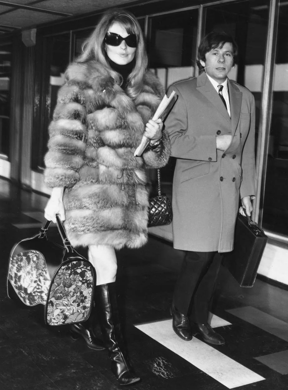 Mulher com casaco de pele, óculos escuros e botas de cano alto, destacando elegância vintage. Bolsa floral adiciona toque artístico. Homem usa casaco clássico com gravata, ressaltando um estilo formal e atemporal. Moda anos 60/70.