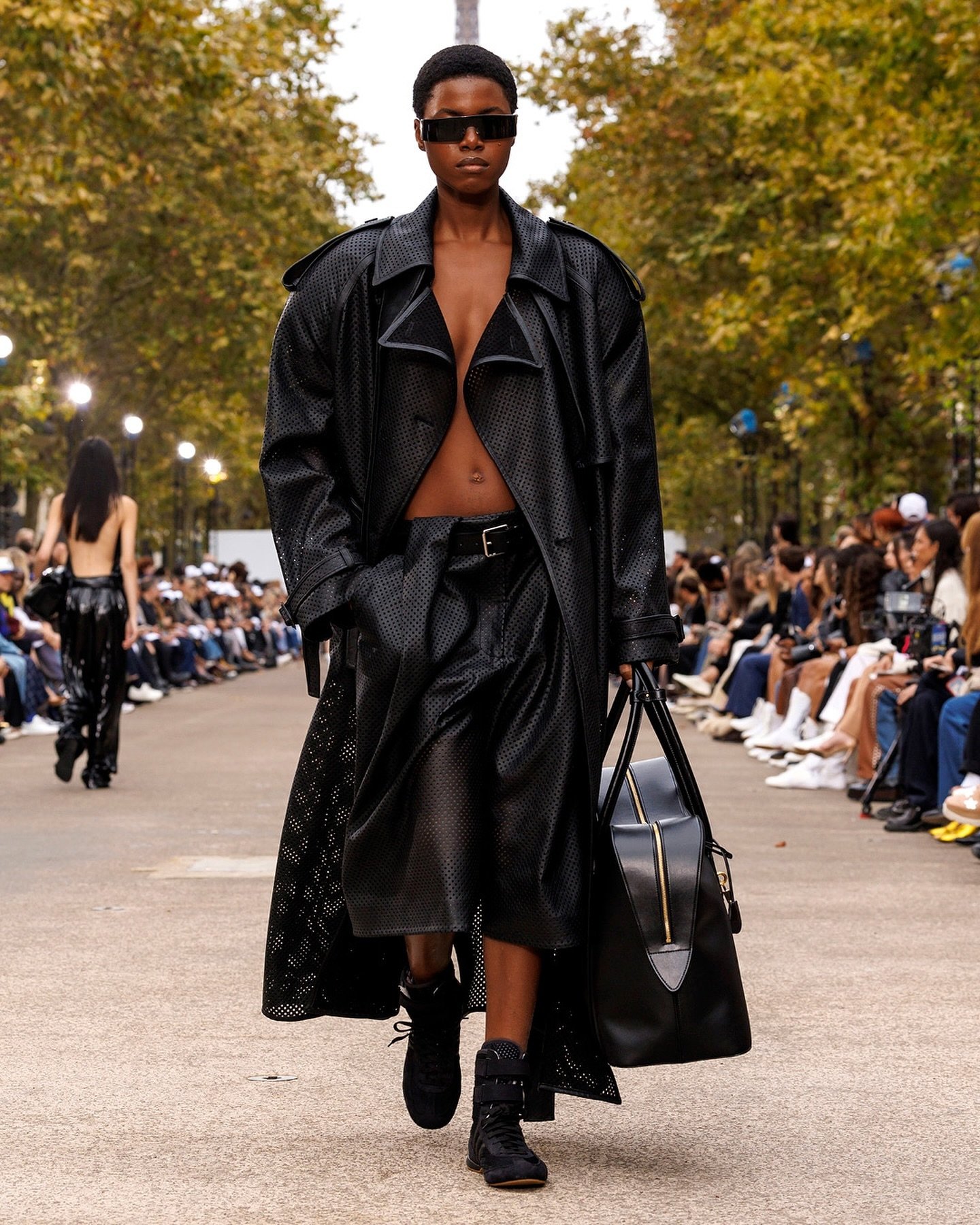 Modelo usando trench coat preto e óculos escuros, caminhando em passarela. Usa botas estilo boxe e carrega bolsa preta.