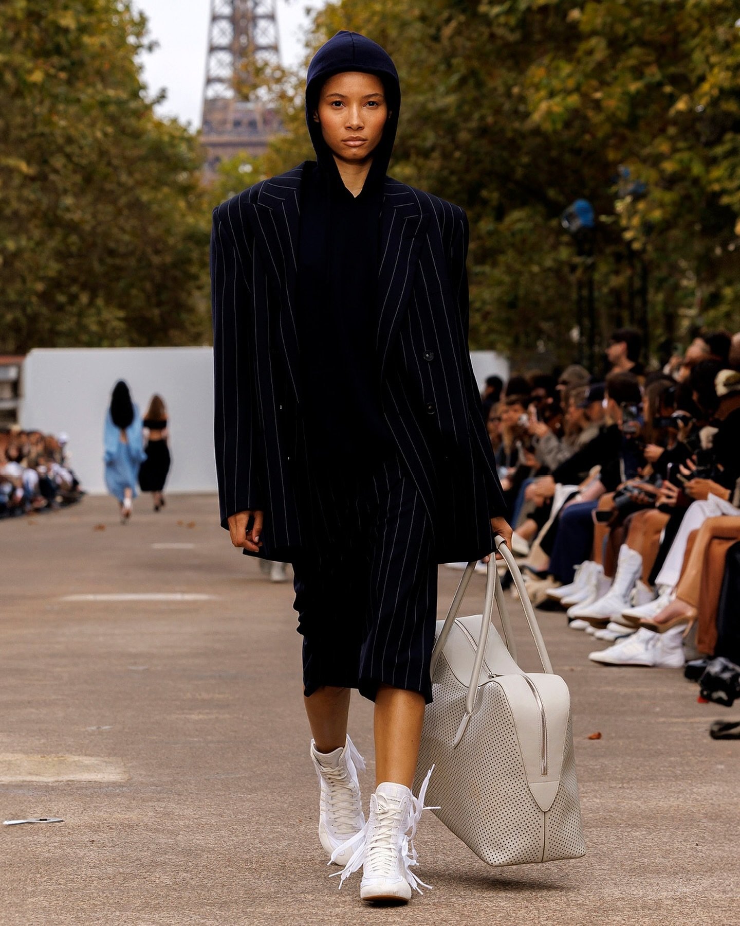 Modelo em blazer risca de giz e capuz combina com botas boxe brancas e bolsa grande em desfile ao ar livre.