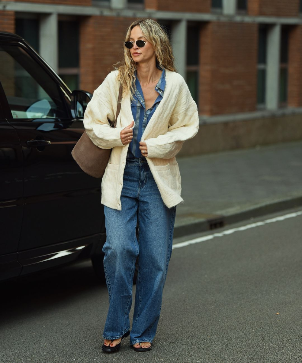 Mulher com cabelo ondulado usando óculos escuros, jeans, camisa de brim e cardigã bege. Look casual e clássico.