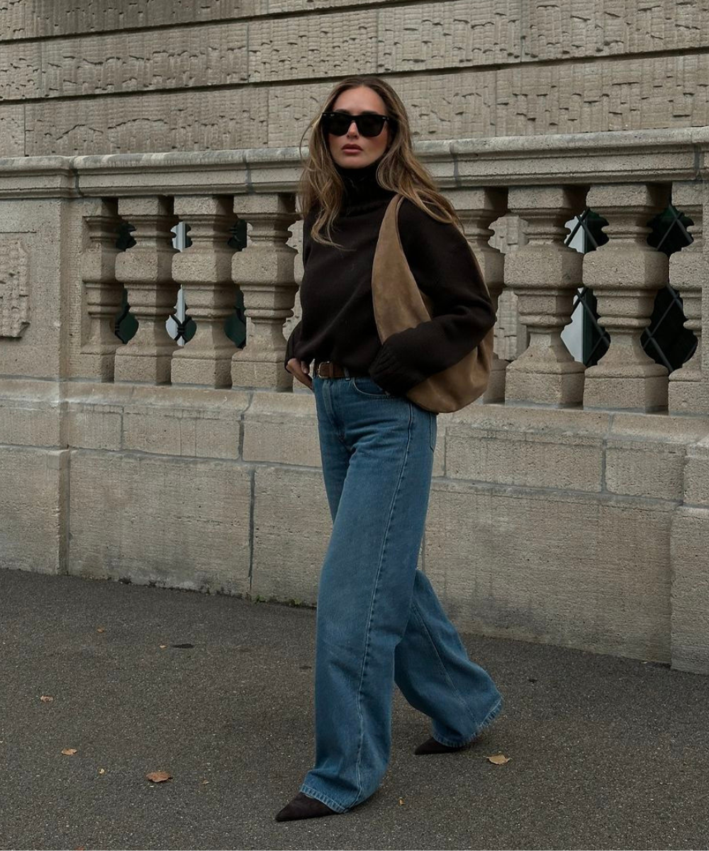 Mulher estilosa com suéter marrom e jeans, usando óculos escuros e bolsa a tiracolo.