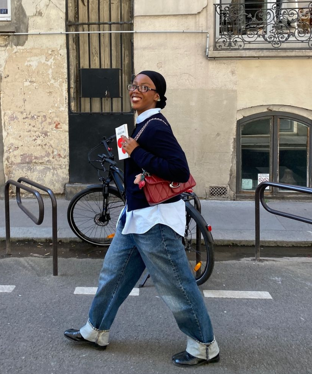 Pessoa caminhando em estilo casual, com jeans e blusa azul. Segura um livro e usa bolsa vermelha. Looks clássicos de todos os tempos.