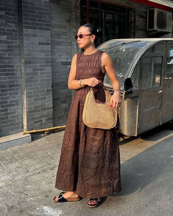 Mulher usando vestido marrom longo e soltinho, sandálias pretas e bolsa de palha. Combinou o look com óculos de sol e acessórios minimalistas. Estilo casual chic, perfeito para dias quentes, misturando elementos naturais e tons neutros.