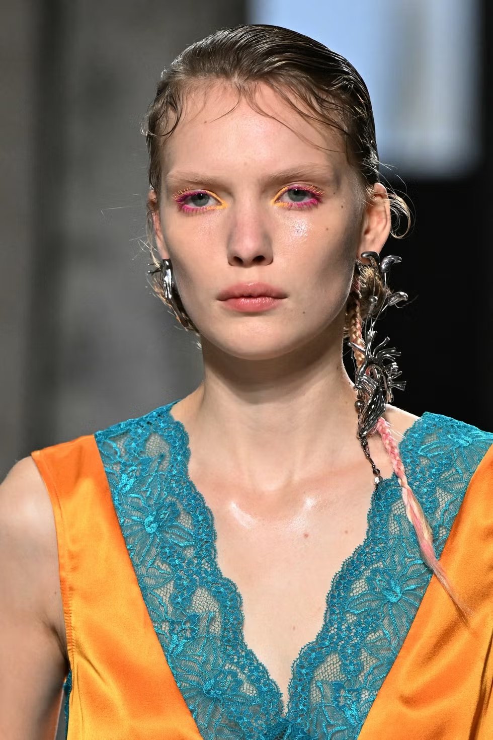 Modelo com vestido de cetim laranja com detalhes em renda azul e maquiagem vibrante nos olhos. O penteado inclui tranças adornadas com acessórios metálicos, destacando uma tendência moderna e futurista na moda. tendências de maquiagem do verão