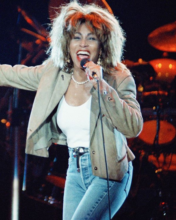 Cantora com cabelo volumoso dos anos 80, usando jaqueta e jeans, canta no palco.