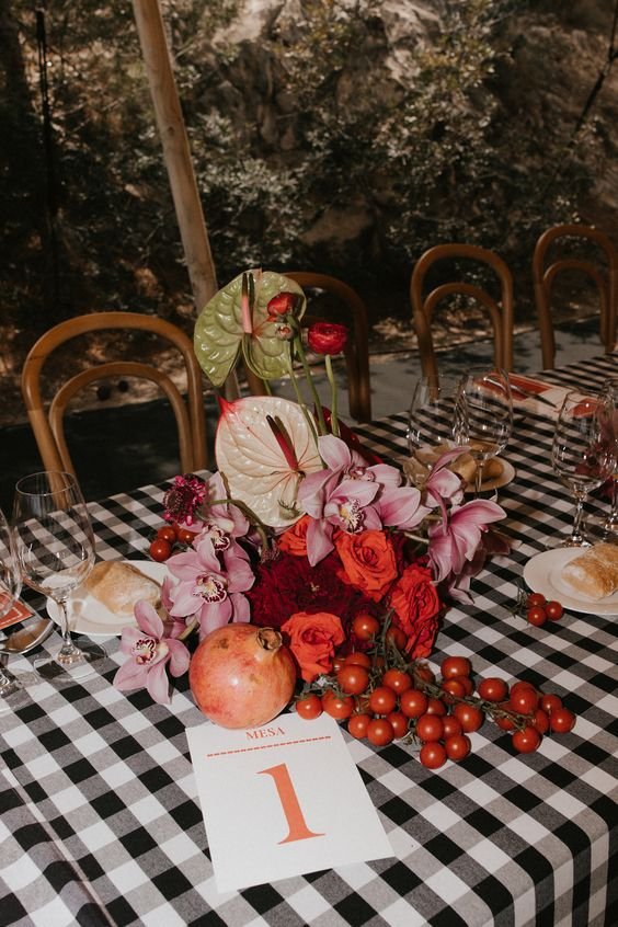 Mesa decorada com estampa xadrez preto e branco, flores vibrantes e frutas como romãs e tomates, criando uma estética rústica e elegante. Tendência de decoração boho-chic, evidenciando contraste entre natural e sofisticado.