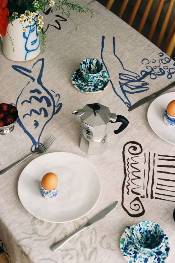 Mesa de café da manhã com toalha estampada, estilo abstrato em azul. Louça com padrão marmorizado em azul e branco, cafeteira italiana e ovos cozidos. Decoração casual e artística, tendência minimalista e boêmia.