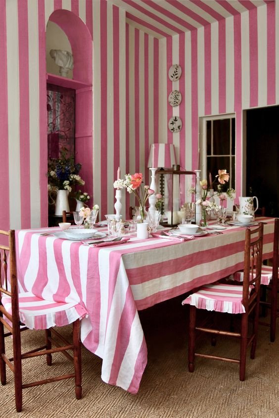 Sala de jantar com decoração vintage e tendência de listras rosa e branco. O ambiente destaca-se pelo uso harmonioso de padrões listrados na toalha de mesa e paredes, criando uma estética retrô-charmosa e acolhedora.