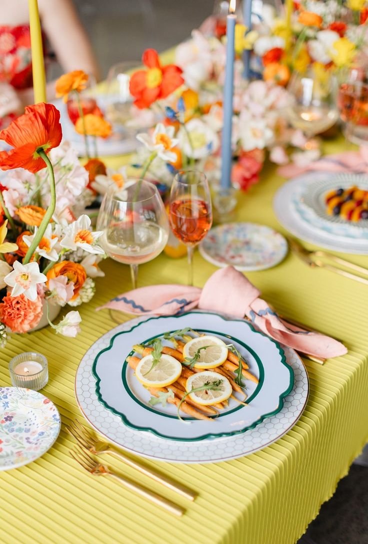 Mesa decorada com flores vibrantes e louça colorida. Toalha amarela e guardanapos rosa destacam-se. Estilo elegante e moderno, ideal para eventos sofisticados e celebrações ao ar livre, seguindo tendências de mesas temáticas e decoração floral.