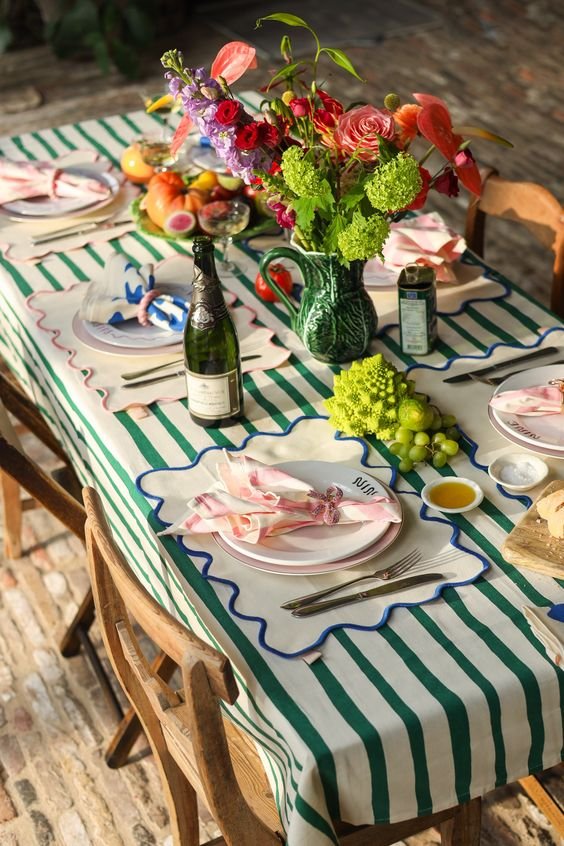 Mesa minimalista e colorida com toalha listrada verde-e-branco. Toalhas e guardanapos com bordas onduladas e vasos de flores vibrantes complementam o visual elegante e casual. Tendência em decoração de mesa para eventos ao ar livre.