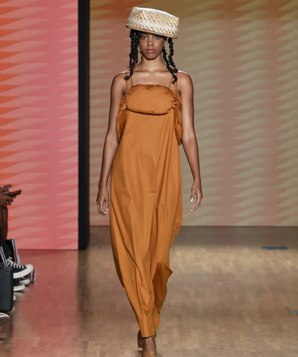 Modelo em desfile SPFW N58 com vestido laranja amplo e chapéu de palha, refletindo tendências contemporâneas.