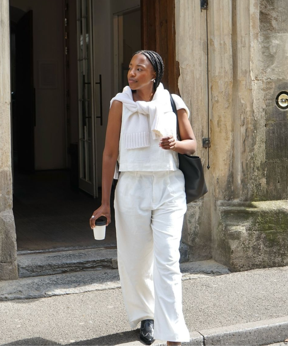 Mulher com look casual e minimalista, usando conjunto branco de calça e blusa. Suéter branco amarrado nos ombros adiciona sofisticação. Bolsa preta e sapatos escuros completam o visual, tendência elegante e prática para o dia a dia urbano.