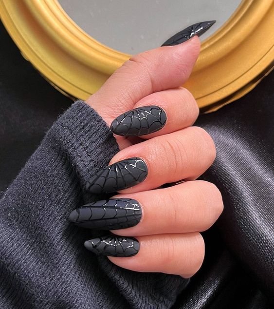 Unhas de halloween longas e pretas com design de teia de aranha, tendência gótica em nail art. A mão veste um suéter cinza escuro, destacando um estilo alternativo e moderno, popular nas tendências de moda outono-inverno.