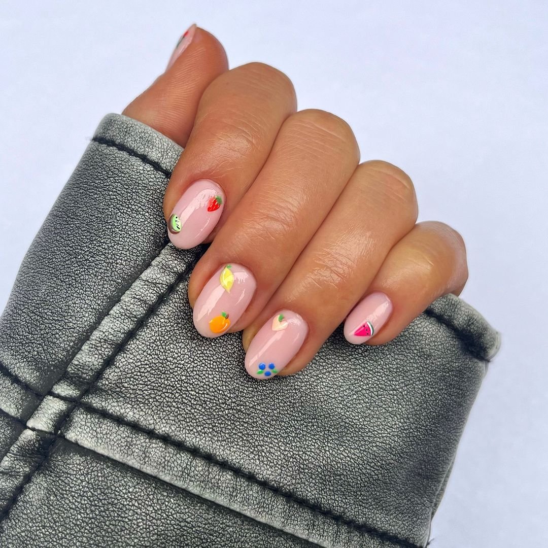 Unhas decoradas com arte de frutas em tons pastel sobre base nude, combinando com uma jaqueta de textura metalizada. Estilo moderno e divertido, destacando a tendência de nail art detalhada. cores de esmalte