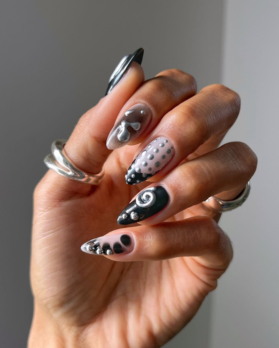 Unhas artísticas em preto, cinza e prata, trazendo tendência moderna com detalhes geométricos e metálicos. Estilo contemporâneo e audacioso, destacado por anéis prateados minimalistas. cores de esmalte