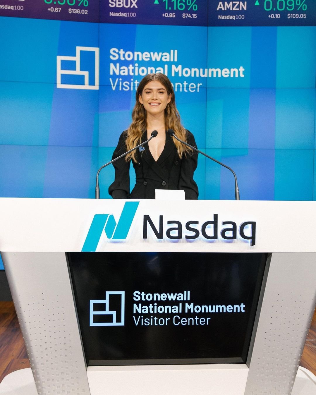 Valentina Sampaio veste blazer preto no pódio da Nasdaq, com painel do Stonewall National Monument ao fundo.