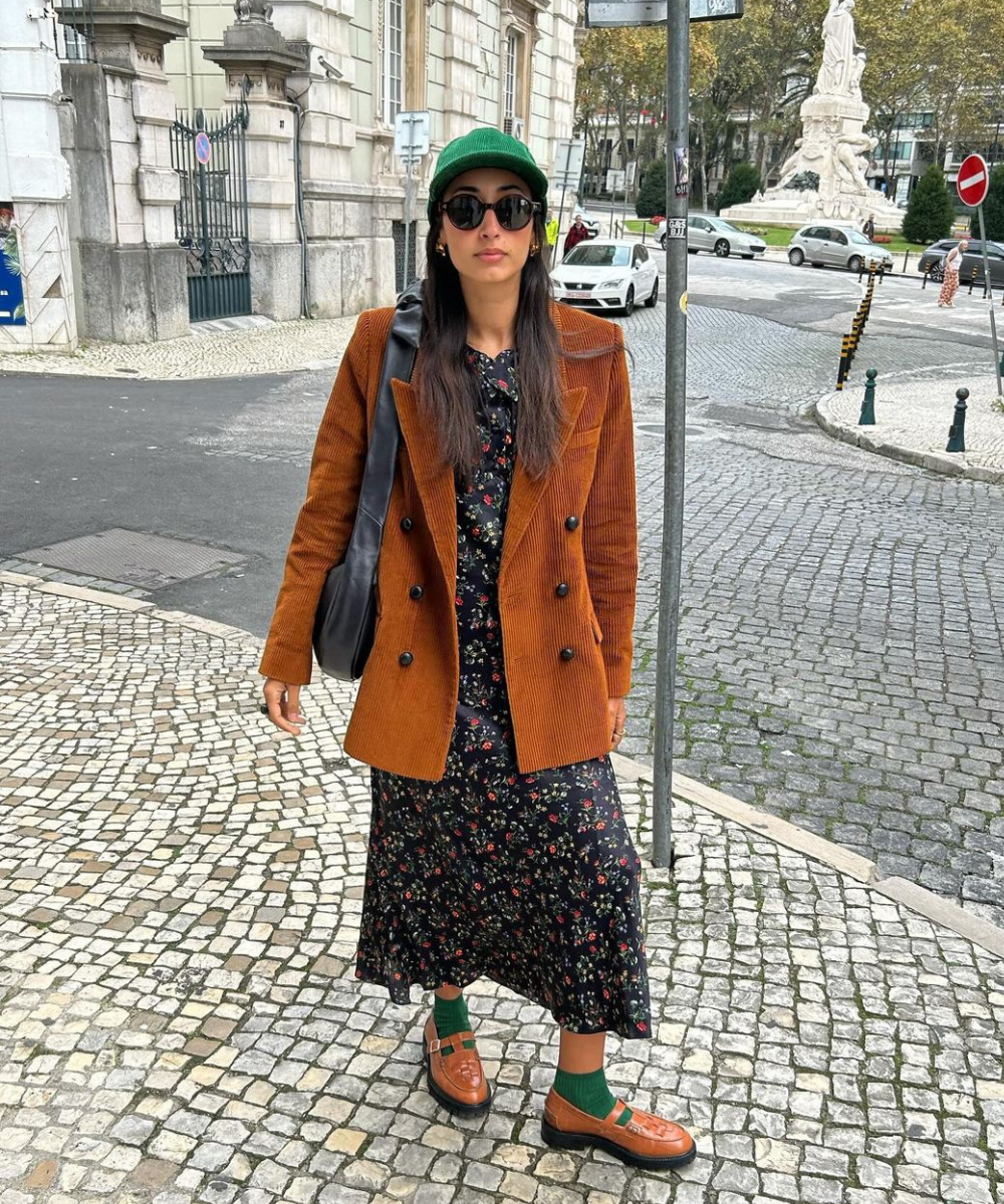 Pessoa com blazer marrom sobre vestido estampado, boné verde e sapatos marrons, caminhando por uma calçada de pedras.