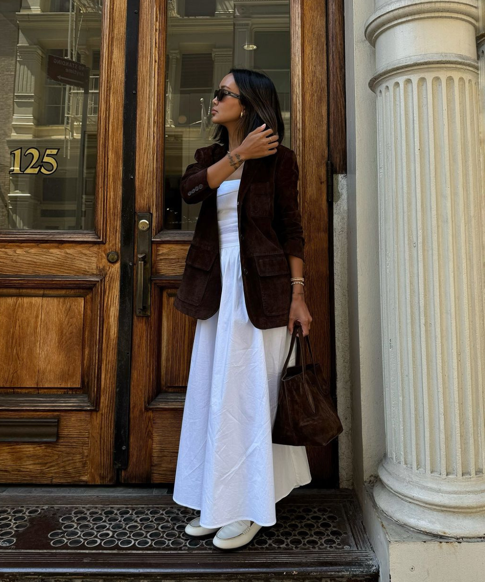 Mulher em pé diante de porta de madeira, usa blazer marrom sobre vestido branco, segurando bolsa combinando.