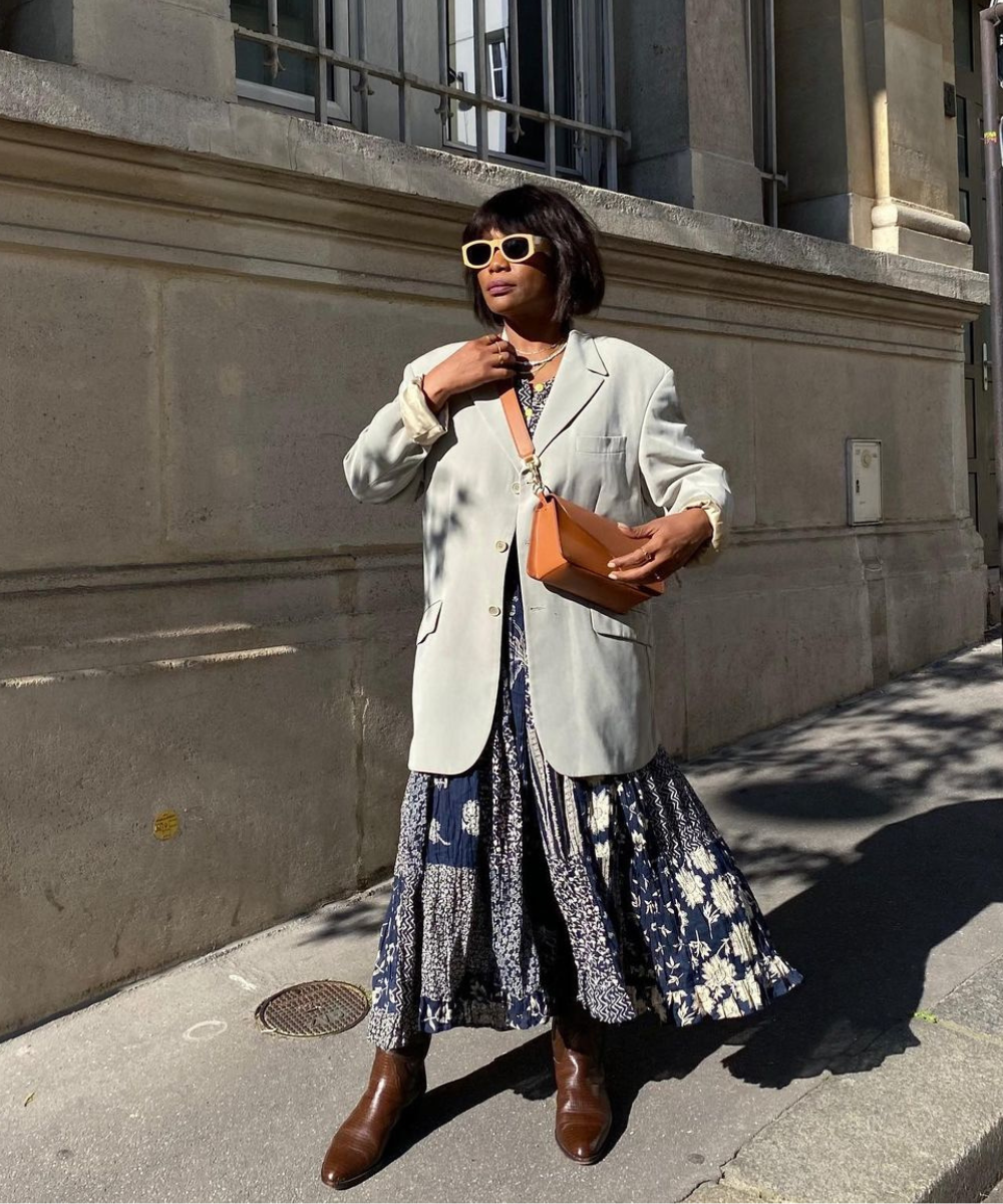 Pessoa com vestido e blazer cinza, segurando bolsa e usando óculos de sol.