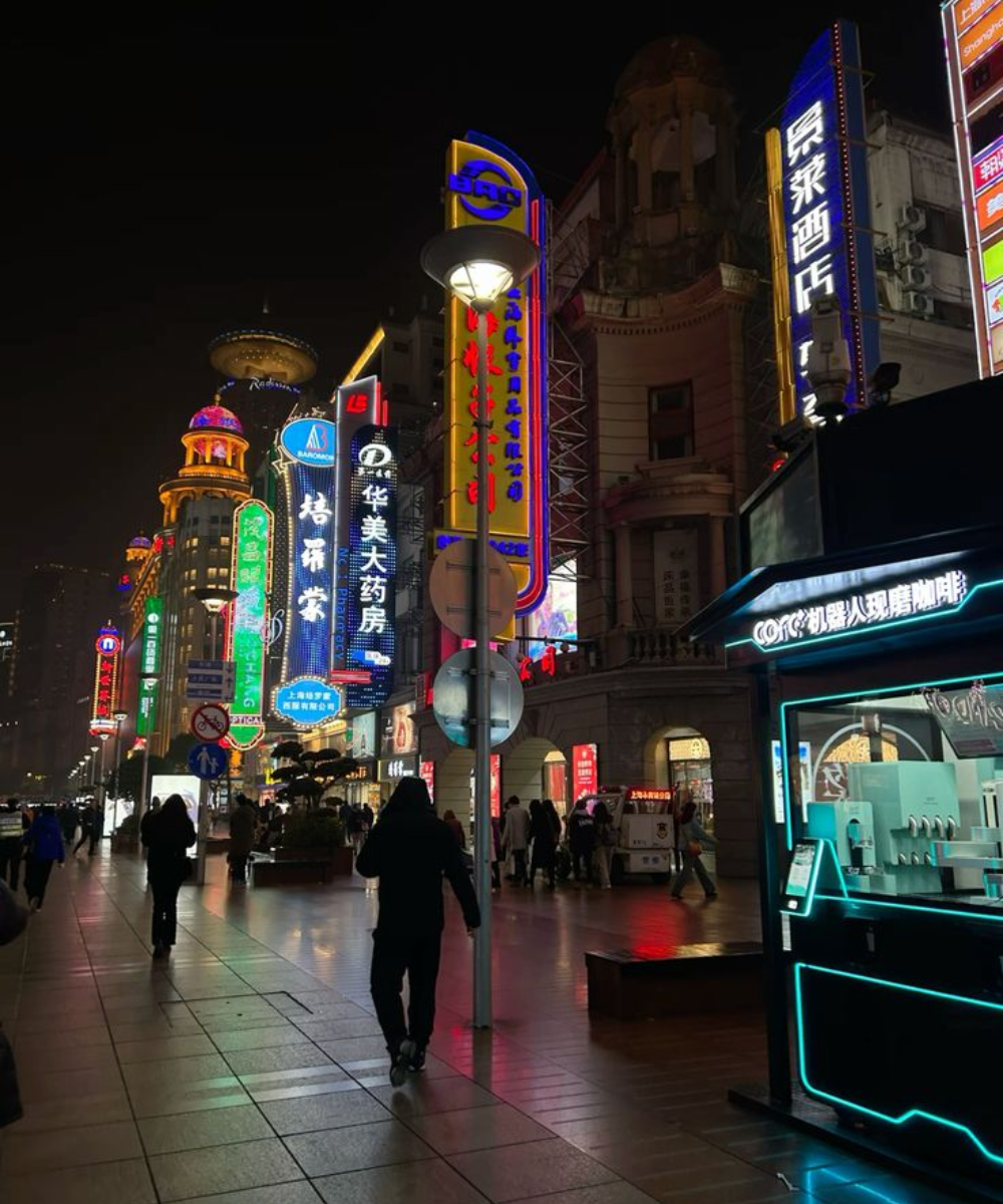 Rua movimentada à noite com destaque para um estilo urbano. Pessoas vestem roupas casuais escuras, como jaquetas e calças estilosas. As luzes de neon adicionam um toque moderno e vibrante, refletindo tendências de moda urbana contemporânea.