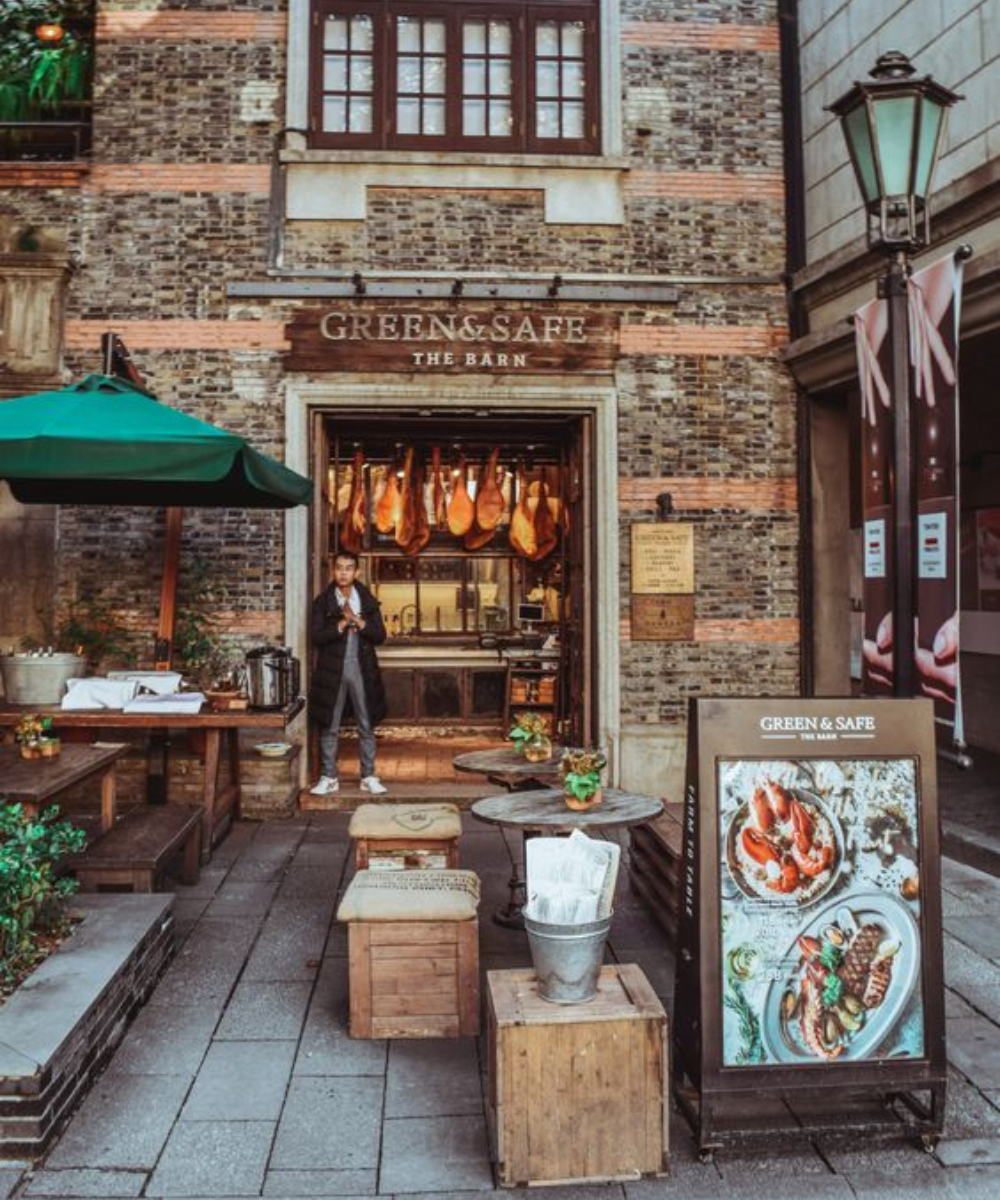 Entrada de café com fachada rústica. Homem veste casaco longo preto, calça jeans e tênis branco, estilo casual urbano. Ambiente combina elementos rústicos e modernos, com mesas de madeira e decoração vintage. Tendência cozy e descontraída.