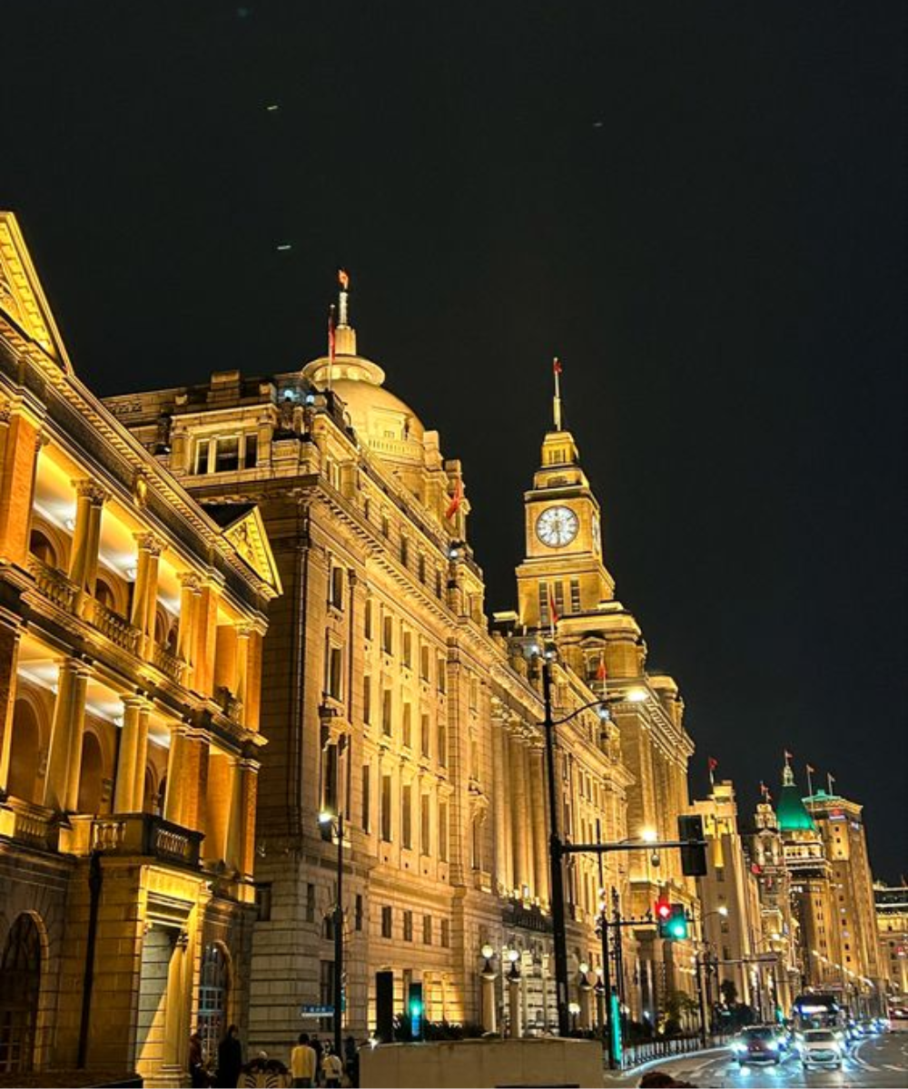 Foto noturna de rua iluminada com arquitetura clássica e imponente. Não há pessoas visíveis para descrever modo de vestir ou tendências de moda. A imagem destaca a beleza das construções históricas sob luzes noturnas.