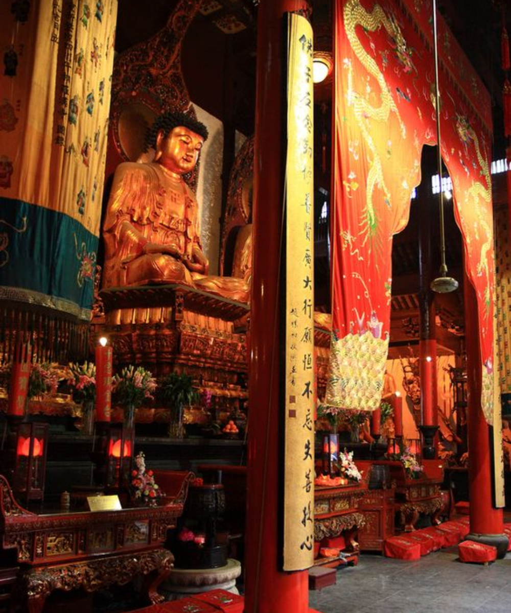 Imagem de um templo com estátua ornamentada usando vestes tradicionais douradas e detalhadas. As tapeçarias ricas em vermelho e dourado destacam a elegância e tradição do local, refletindo tendências de moda histórica asiática.