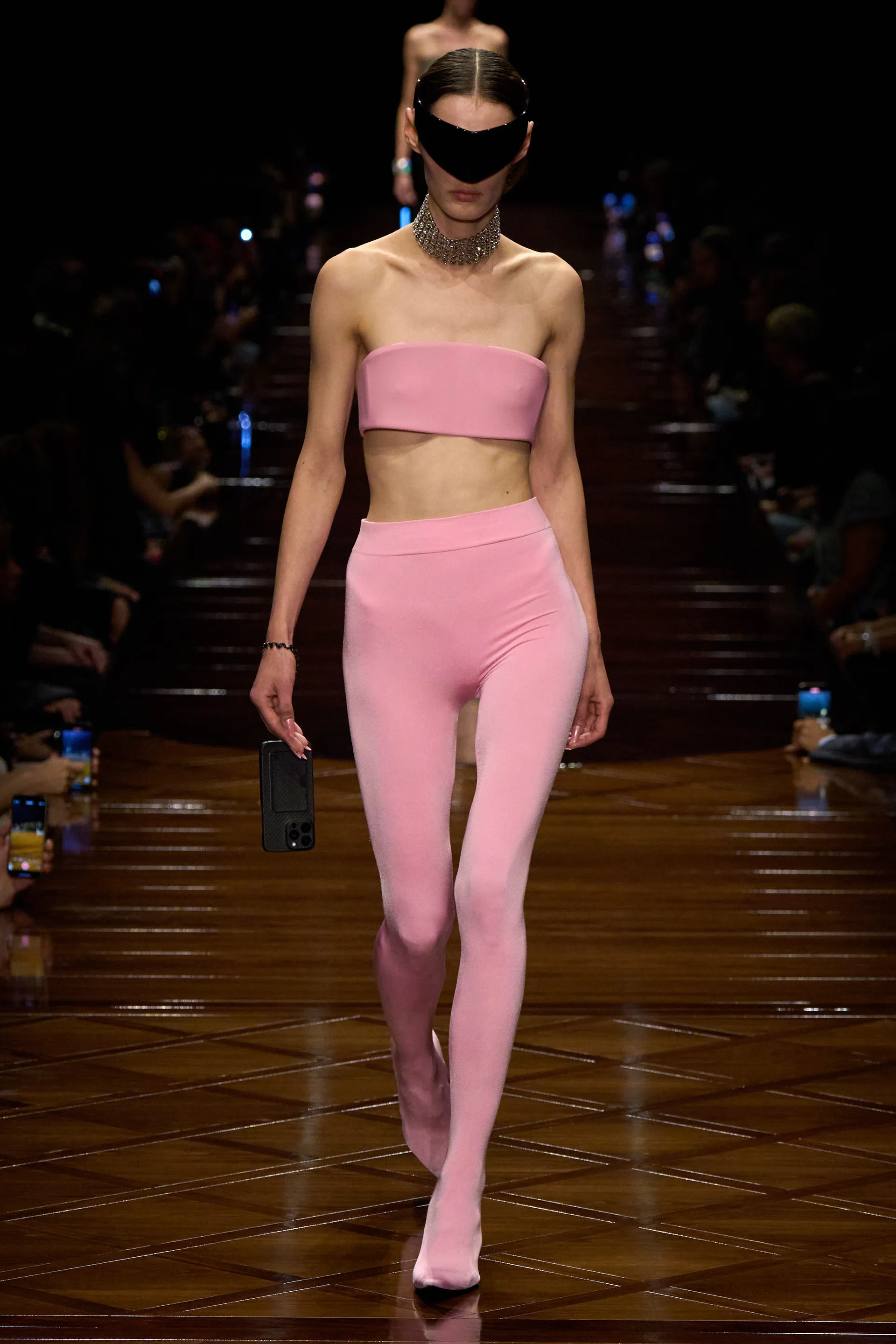 Modelo de top e calça rosa com máscara no rosto em desfile, refletindo tendências das cores do verão 2025.