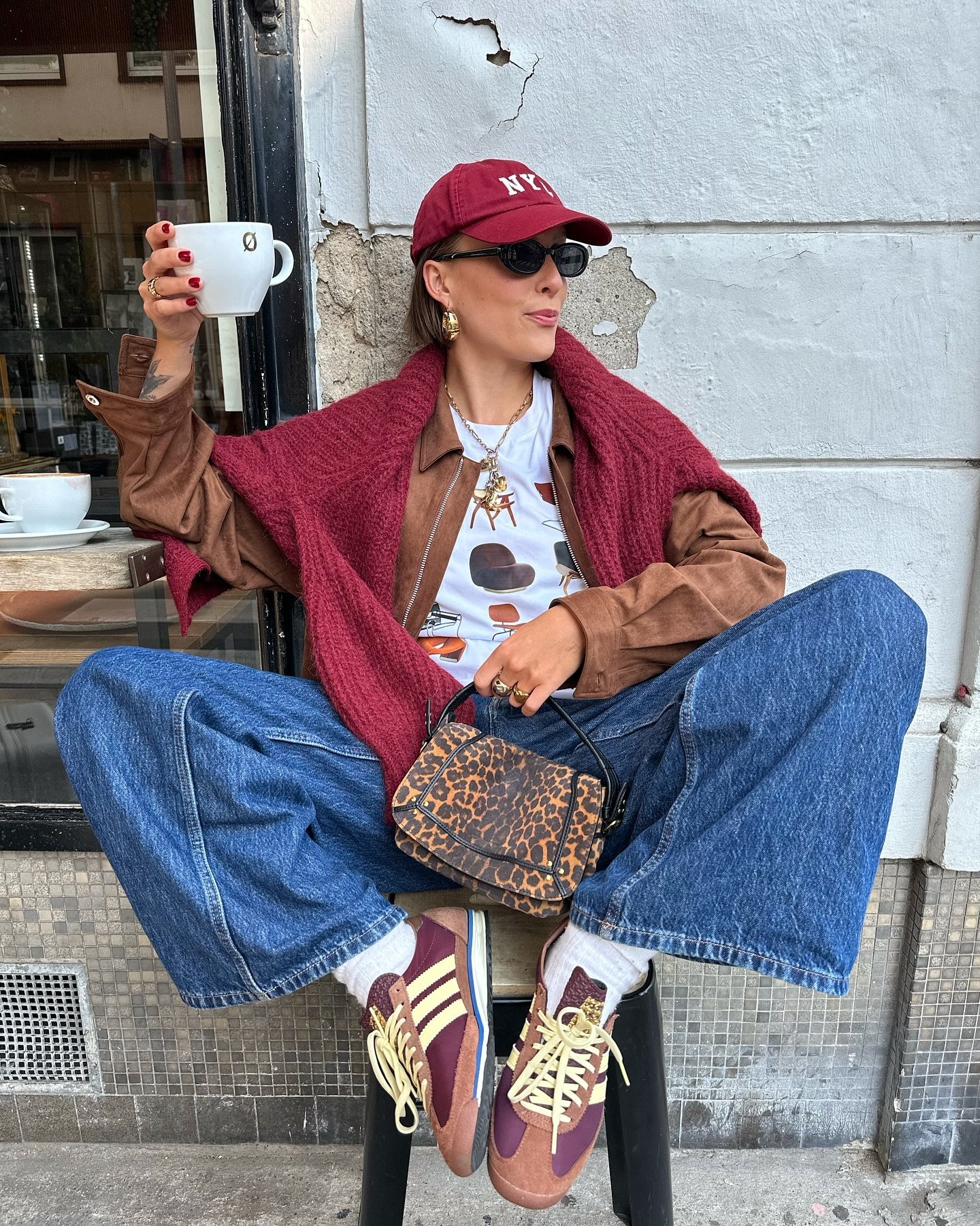 Pessoa sentada em um banco, segurando uma xícara de café, com roupa casual e bolsa animal print. Atmosfera descontraída.